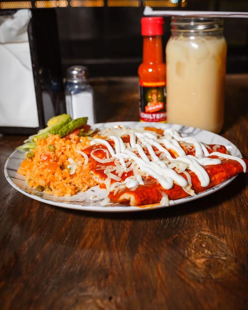 A plate of enchiladas with rice and salad, hot sauce, and a drink on a wooden table.