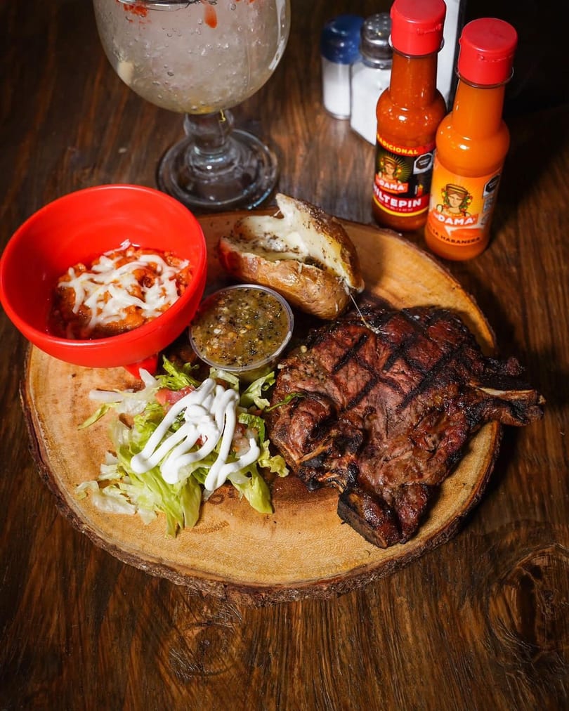 Grilled steak with baked potato, beans, salad, salsa, and hot sauce on a wooden board.