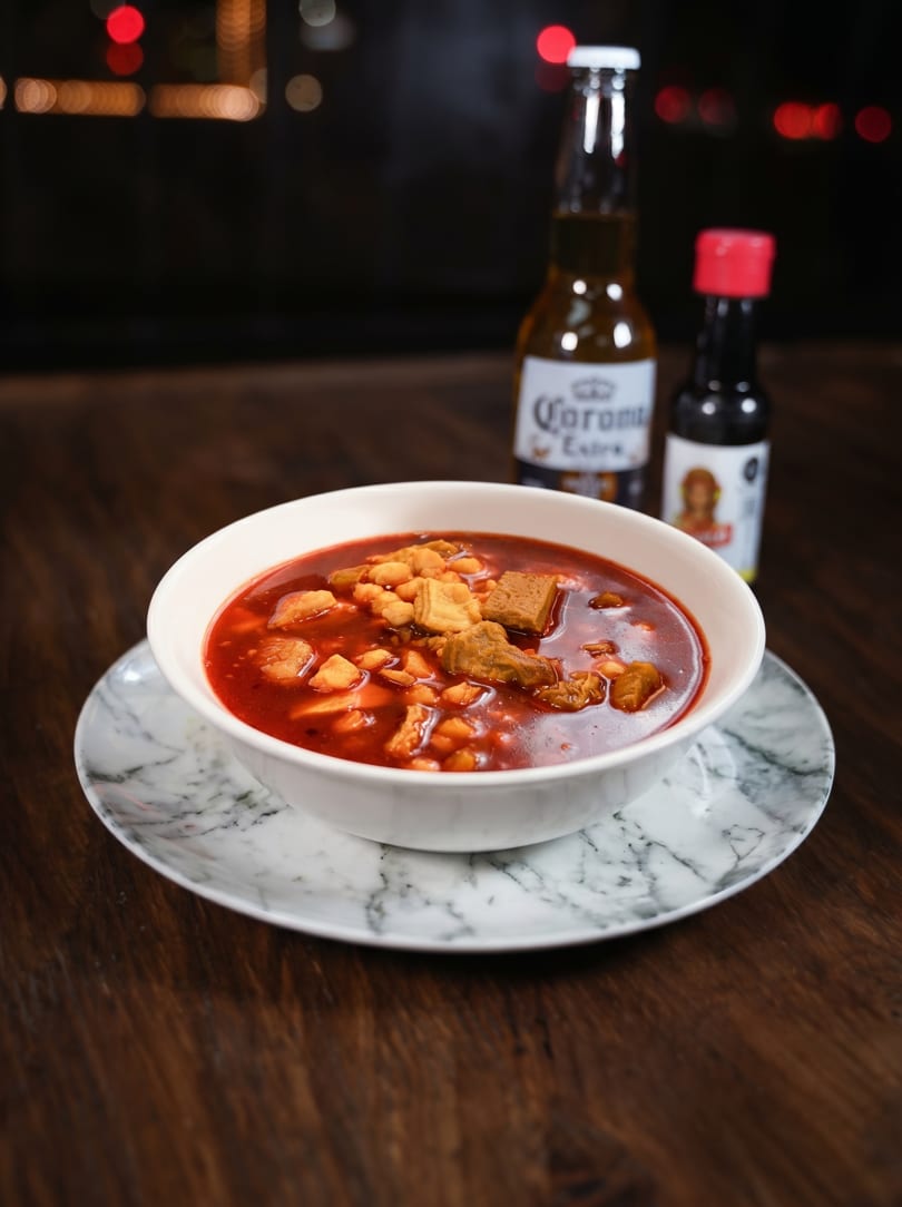 A bowl of red pozole with meat and hominy, a Corona beer, and hot sauce.