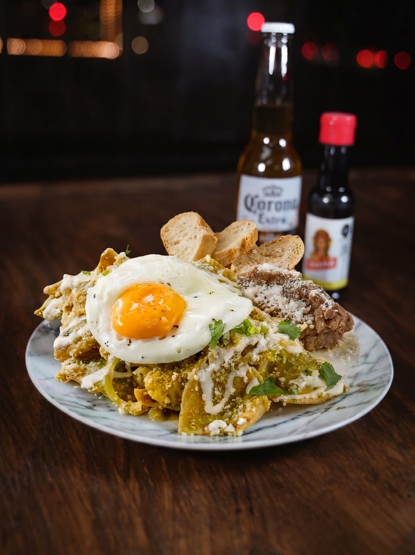 Chilaquiles with a fried egg, refried beans, and bread, served with a Corona beer and hot sauce.