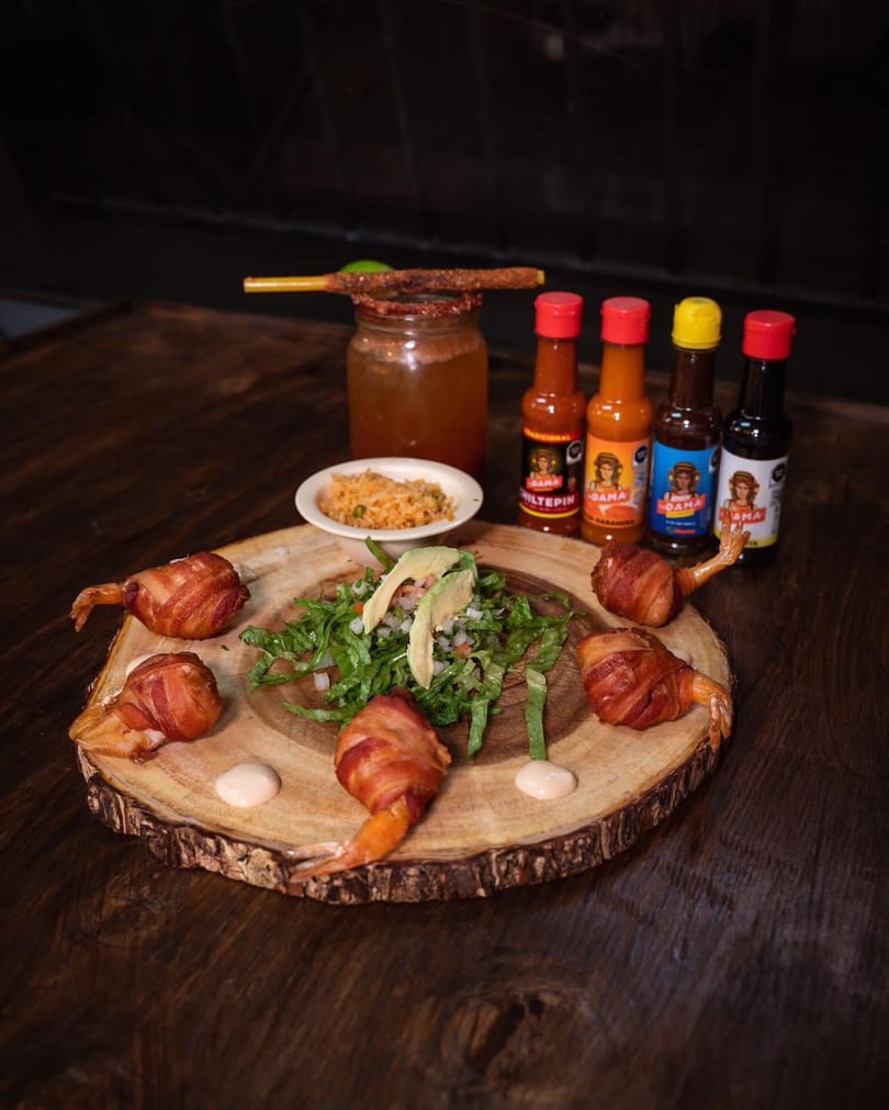 Bacon-wrapped shrimp, salad, rice, hot sauces, and a michelada on a wooden board.